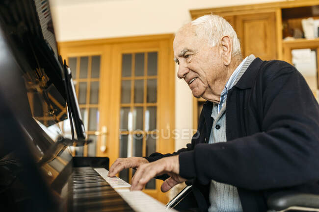 Hombre mayor sonriente sentado en silla de ruedas tocando el piano en casa - foto de stock