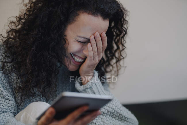 Mujer sentada en casa, usando tableta digital, riendo - foto de stock