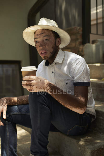 Portrait of young man with vitiligo wearing a hat, holding a takeaway coffee sitting on the stairs — Stock Photo