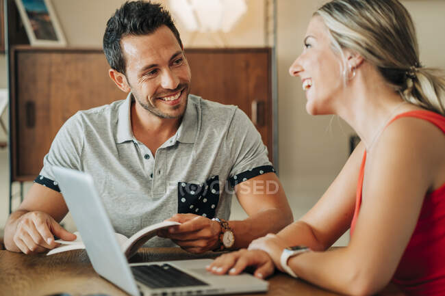 Glückliches Paar sitzt mit Laptop und Buch am Tisch — Stockfoto