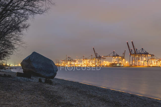 Allemagne, Hambourg, Alter Schwede rocher sur la plage au bord de la rivière au crépuscule avec des silhouettes de grues portuaires en arrière-plan — Photo de stock