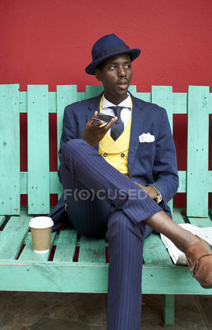 Молодий бізнесмен в старомодному костюмі і капелюсі сидить на зеленій лавці, використовуючи свій телефон — стокове фото