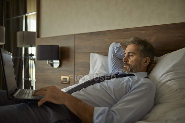 Businessman lying on bed in hotel room using laptop — Stock Photo