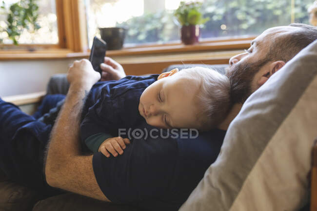 Man checking his phone while his little baby son is sleeping on the sofa — Stock Photo