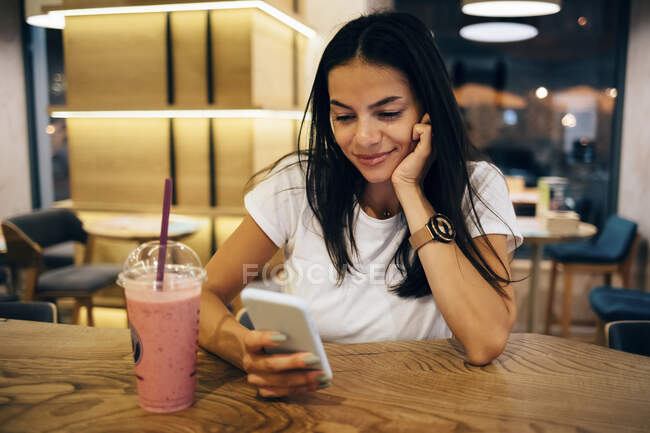 Black-haired woman drinking a smoothie and using smartphone in cafe — Stock Photo