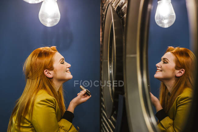 Улыбающаяся рыжеволосая молодая женщина смотрит на свое зеркальное отражение помадой — стоковое фото