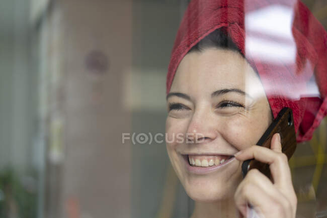 Retrato de mujer feliz con la cabeza envuelta en una toalla hablando por teléfono detrás del cristal de la ventana - foto de stock