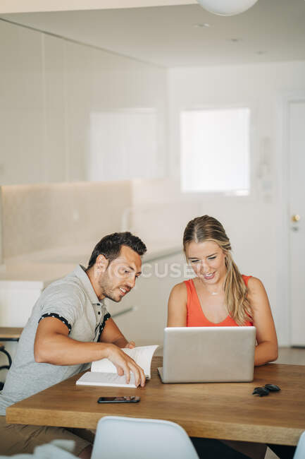 Glückliches Paar sitzt mit Laptop und Buch am Tisch — Stockfoto