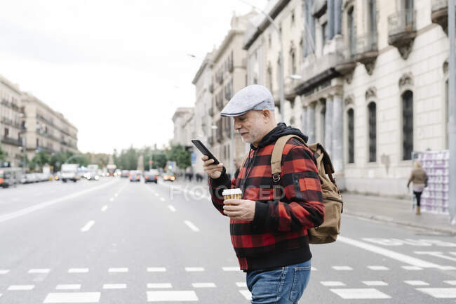 Senior man with zaino and coffee to go looking at cell phone while crossing the street, Barcellona, Spagna — Foto stock