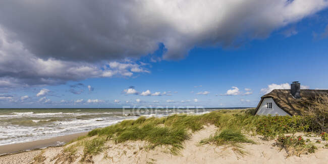 Alemanha, Mecklemburgo-Pomerânia Ocidental, Ahrenshoop, Nuvens sobre casa com telhado de palha em pé na praia costeira arenosa do Mar Báltico — Fotografia de Stock