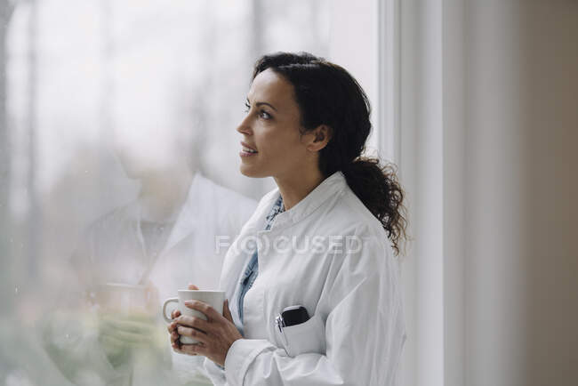 Жінка-лікар дивиться з вікна, тримає чашку кави — стокове фото