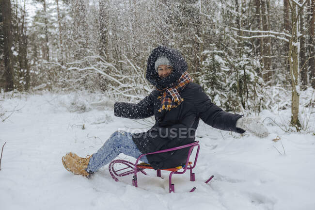 Усміхнена жінка сидить на санях, кидаючи сніг у повітря в зимовий ліс. — стокове фото