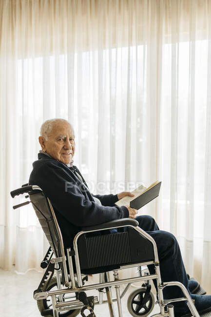 Retrato de un hombre mayor sonriente sentado en silla de ruedas con un libro - foto de stock