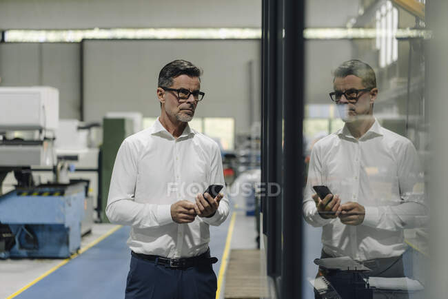 Homme d'affaires mature avec téléphone portable dans une usine reflétée dans la vitre — Photo de stock