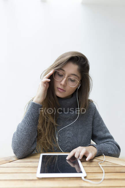 Portrait of young woman sitting at wooden table using earphones and digital tablet — Stock Photo