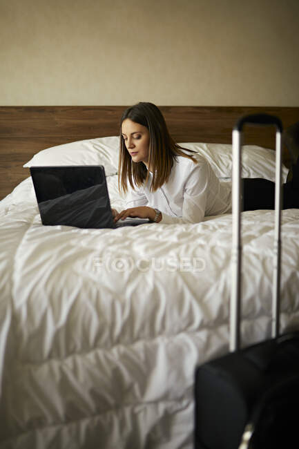 Mujer de negocios acostada en la cama en la habitación de hotel utilizando el ordenador portátil - foto de stock