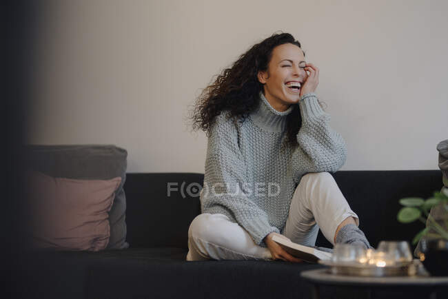 Mujer sentada en el sofá riendo, leyendo un libro - foto de stock