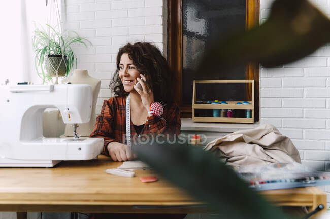 Mujer en el teléfono sentada en la mesa con máquina de coser - foto de stock