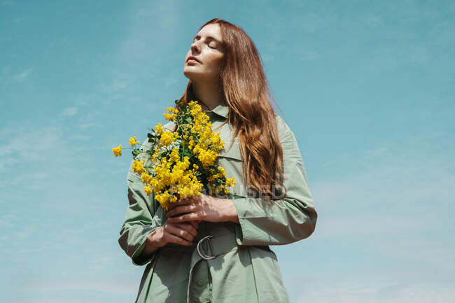 Portrait of redheaded young woman with eyes closed standing against sky holding bunch of yellow flowers — Stock Photo