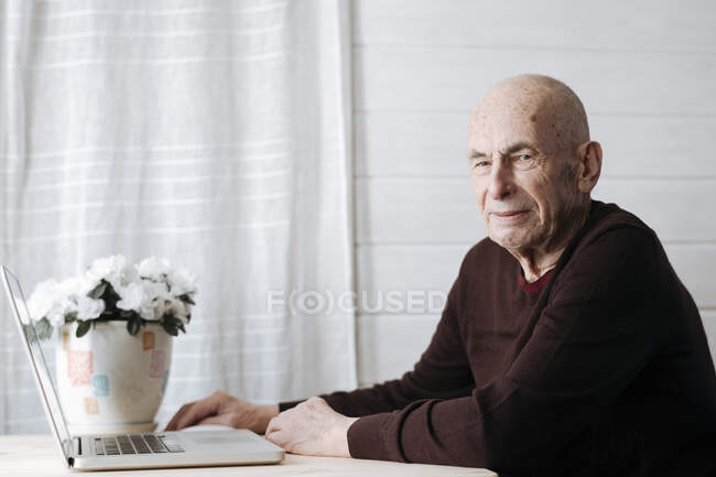 Retrato del anciano sonriente sentado en la mesa con el ordenador portátil - foto de stock