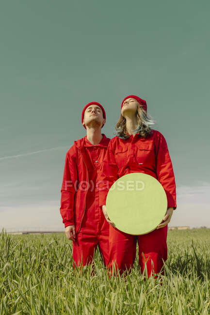 Pareja joven vestida con monos rojos de pie en un campo con círculo verde - foto de stock