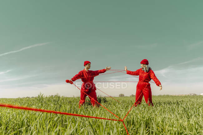 Young couple wearing red overalls and hats performing on a field with red string — Stock Photo