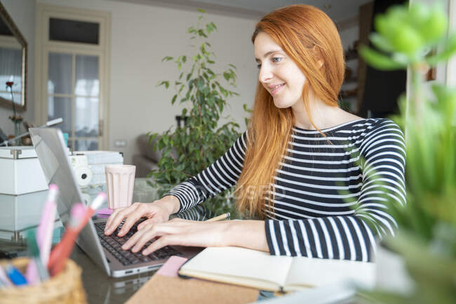 Mujer joven sonriente usando el ordenador portátil en el escritorio en casa - foto de stock