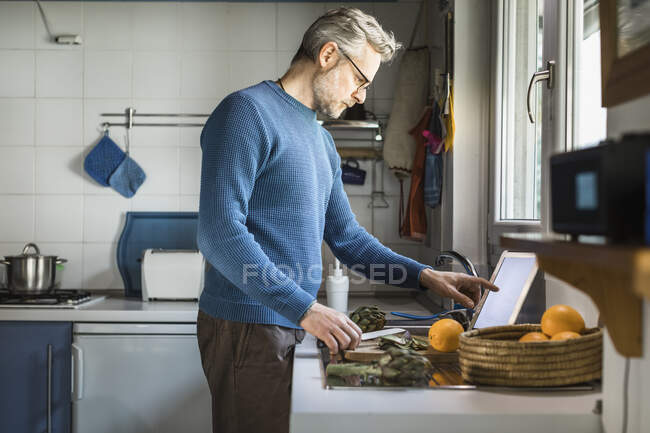 Hombre maduro preparando alcachofas en su cocina usando tableta digital - foto de stock
