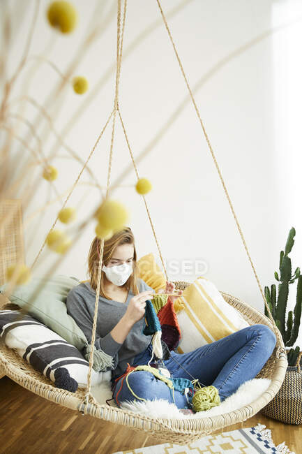 Блондинка в маске FFP2 и вязании сидит в висячем кресле дома — стоковое фото
