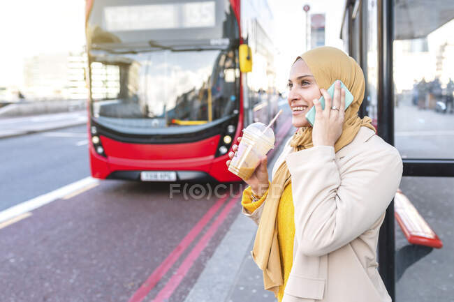 Portrait of smiling young woman on the phone waiting at bus stop — Stock Photo
