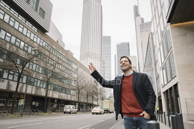 Усміхаючись середньодорослому бізнесмену, який вітає таксі, стоячи на тротуарі в місті, відверті, зародкові — стокове фото