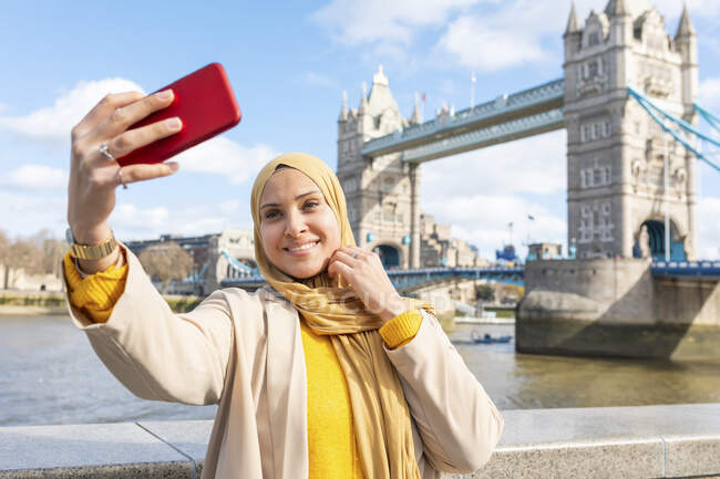 Retrato de una joven sonriente tomando selfie con smartphone frente a Tower Bridge, Londres, Reino Unido - foto de stock