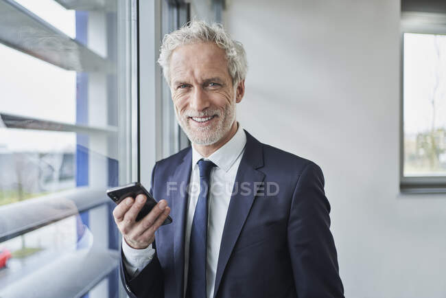 Portrait of smiling businessman holding cell phone at the window — Stock Photo