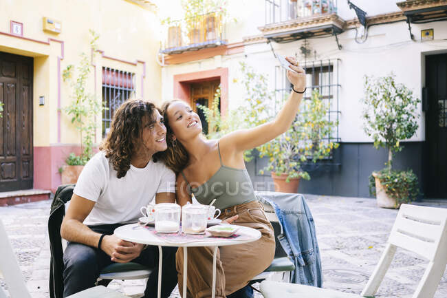 Giovane donna che prende selfie attraverso il telefono cellulare con il fidanzato mentre è seduto al caffè marciapiede, Santa Cruz, Siviglia, Spagna — Foto stock