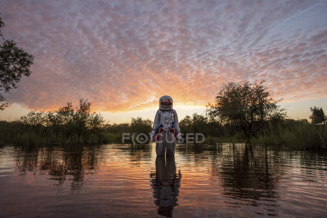 Spacewoman standing in water at sunset — spacesuit, tree - Stock Photo