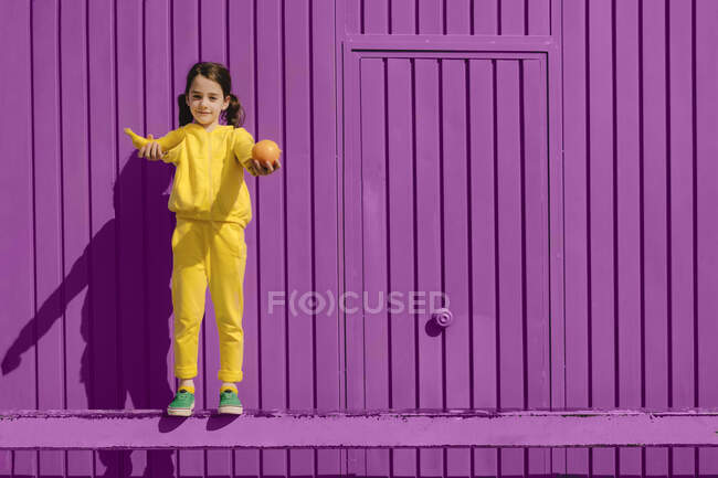 Portrait de petite fille vêtue de jaune debout devant un fond violet offrant des fruits — Photo de stock