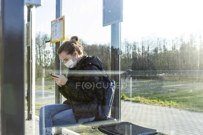 Девушка в маске ждет на автобусной остановке, используя смартфон — стоковое фото