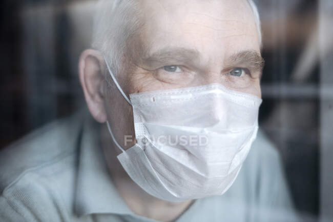 Портрет пожилого человека в маске дома, смотрящего в окно — стоковое фото