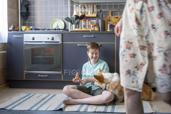 Ragazzo sorridente seduto sul pavimento della cucina con un pezzo di pizza che guarda il cane — Foto stock