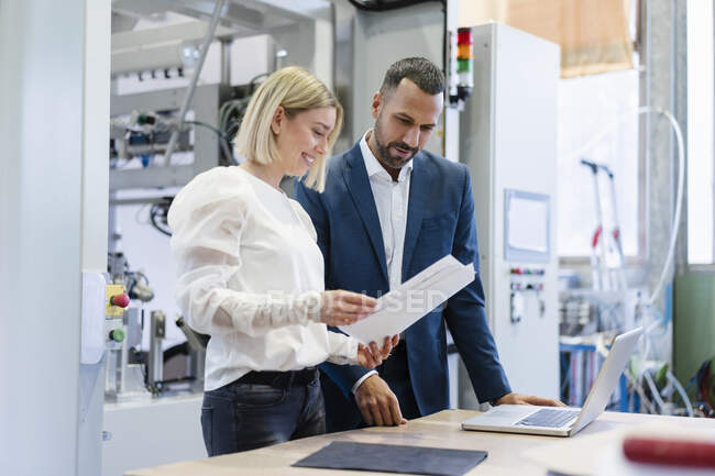 Businessman and young woman with papers and laptop talking in a factory — Stock Photo