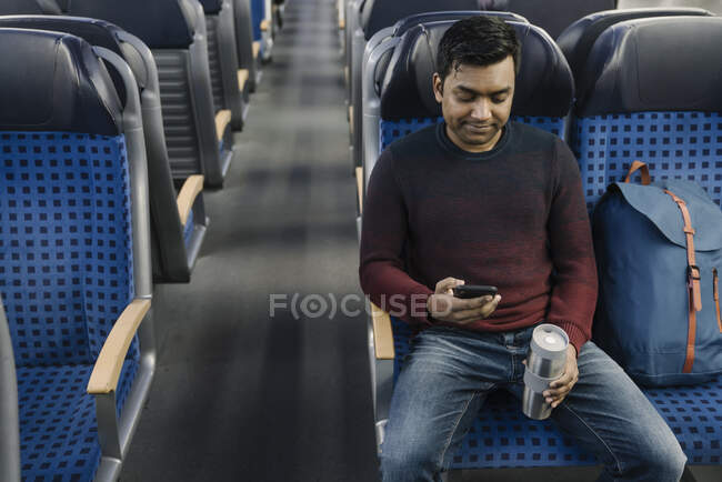 Hombre sosteniendo taza reutilizable y el uso de teléfono inteligente en el tren - foto de stock