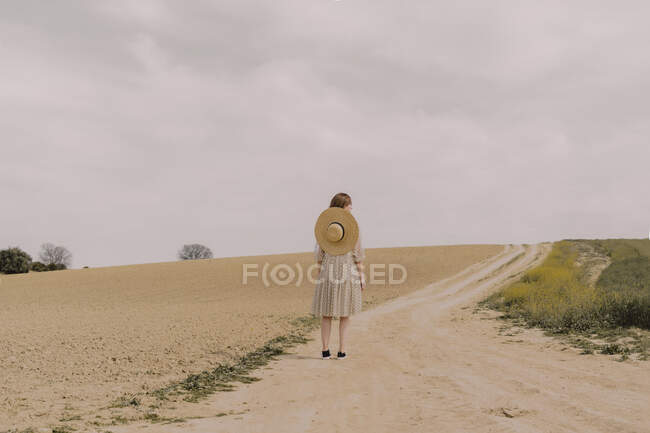 Mulher com chapéu de palha e vestido vintage sozinho em uma estrada de campo remoto no campo — Fotografia de Stock