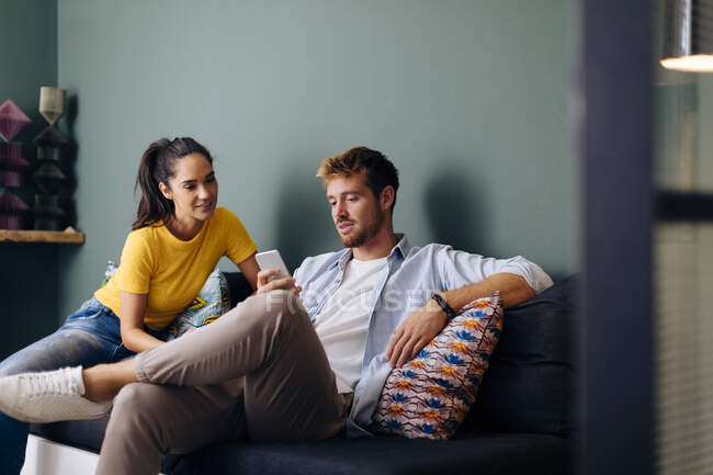 Giovane coppia seduta sul divano a casa guardando lo smartphone — Foto stock
