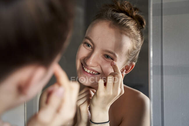 Immagine a specchio della ragazza ridente che applica la crema per il viso in bagno — Foto stock