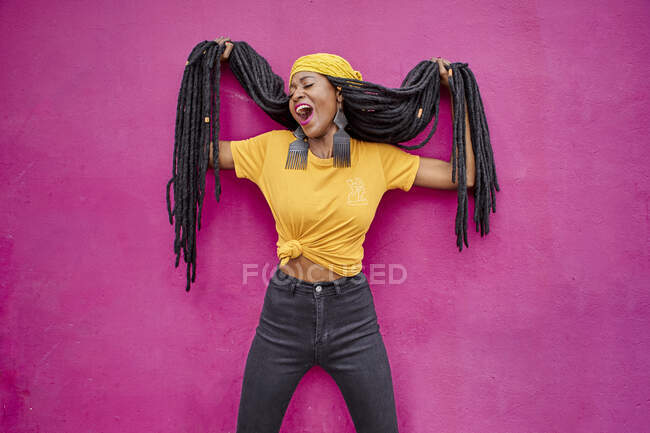 Retrato de mujer con rastas largas sosteniendo su cabello frente a una pared rosa - foto de stock