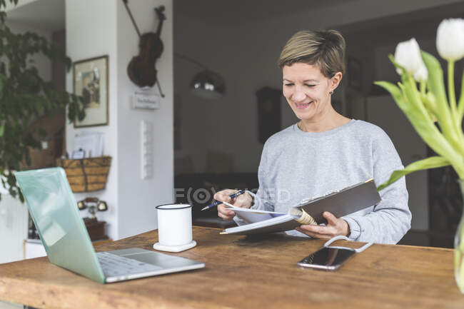 Femme mûre travaillant de la maison, en utilisant un ordinateur portable sur la table avec des fleurs — Photo de stock