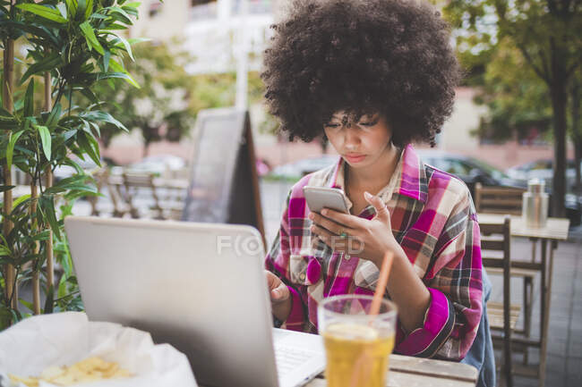 Young woman with afro hairdo using smartphone and laptop at an outdoor cafe in the city — Stock Photo