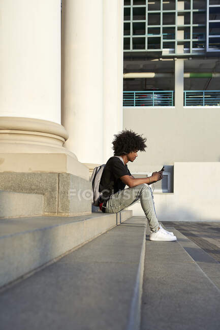 Giovane uomo che ascolta musica e tiene lo smartphone, seduto sui gradini — Foto stock