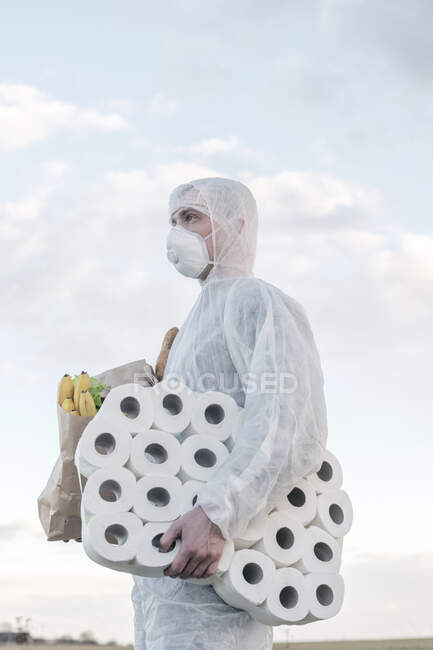 Homem vestindo terno protetor e máscara segurando rolos de banheiro e saco de supermercado ao ar livre — Fotografia de Stock