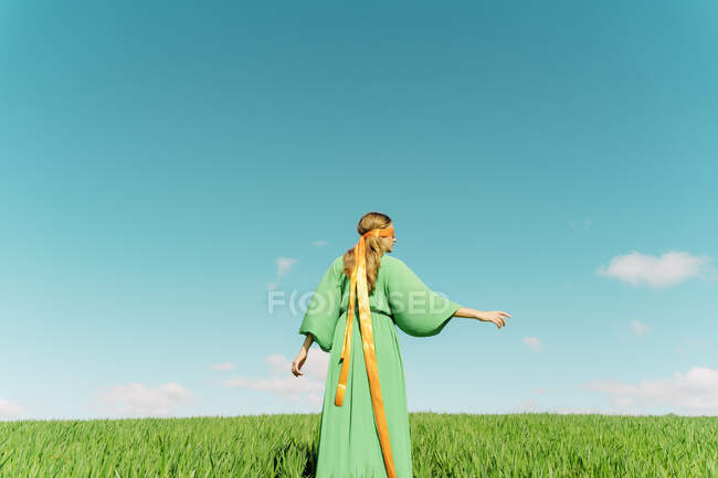 Vista posteriore della giovane donna che indossa un abito verde in piedi in un campo con un nastro intorno alla testa — Foto stock